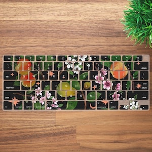 Op de afbeelding: Een zwart toetsenbordhoes met een bloemenontwerp met appels, peren en bloesems. Het ontwerp is op een zwarte achtergrond met groene bladeren en witte bloemen.