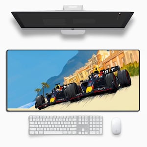 May include: A rectangular mouse pad featuring a vibrant illustration of two Formula 1 race cars speeding along a track. The cars are primarily black, yellow, and red, set against a backdrop of a coastal town and blue sky. A white keyboard and mouse are below.