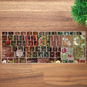 Może przedstawiać: Dekoracyjna nakładka na klawiaturę z kwiatowym i botanicznym wzorem. Klawisze zdobią różne wzory i kolory, w tym czerwony, różowy, zielony i brązowy. Nakładka na klawiaturę jest umieszczona na drewnianej powierzchni.