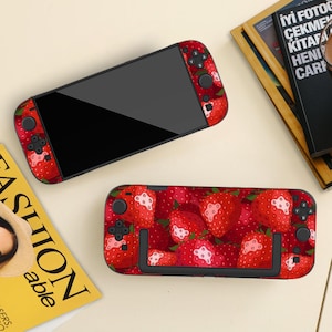 May include: Two black handheld gaming consoles with a vibrant red strawberry pattern. The consoles have black buttons and a black screen. One console is in two parts, and the other is whole. The background is a light beige colour.