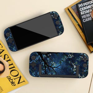 May include: Two handheld gaming consoles with floral designs. One has a dark blue floral pattern, the other a night scene with branches and white flowers. The screens are black. The consoles are on a light surface.