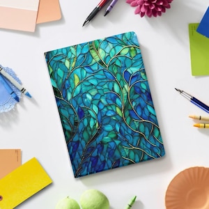 May include: A rectangular journal with a stained glass design featuring blue and green leaves with gold-colored veins. The journal is surrounded by pens, crayons, and other stationery items on a white surface.
