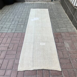 Puede incluir: Una alfombra larga de tejido blanco roto con una textura sutil. La alfombra está colocada en un patio de ladrillo gris y marrón.