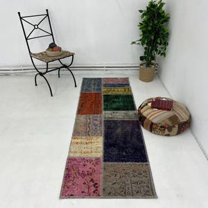 May include: A colorful patchwork runner rug with a variety of patterns and colors, including pink, green, blue, and brown. The rug is laid out on a white floor with a black metal chair and a patterned floor cushion in the background.