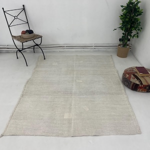 Może przedstawiać: Prostokątny, jasno-beżowy dywan o teksturowanej powierzchni. Dywan leży na białej podłodze. Czarny metalowy krzesło z tkanym siedziskiem i mała roślina doniczkowa znajdują się w tle.