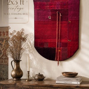 May include: A rectangular vintage wall hanging rug in shades of deep red and burgundy, featuring a patchwork design with a rounded bottom edge and two long braided cords hanging from the center. Text on the image reads 2x3 ft vintage WALL HANGING RUG.