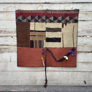 May include: A rectangular woven wall hanging featuring a top band of dark red and geometric black and white patterns. The centre section consists of beige and brown striped panels, while the bottom is a solid rust-coloured textile with a braided cord.