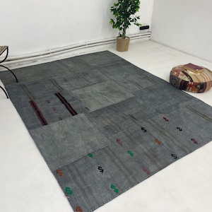 Puede incluir: Una alfombra gris de patchwork con una variedad de texturas y patrones. La alfombra está hecha de diferentes piezas de tela cosidas juntas. La alfombra está en una habitación con paredes blancas y una mesa pequeña con una silla.