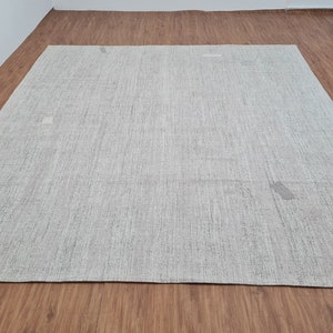 May include: A large, rectangular, light grey rug with a textured surface. The rug is placed on a wooden floor, and a stack of folded textiles is visible in the background. The rug's neutral colour and simple design make it suitable for various interior styles.