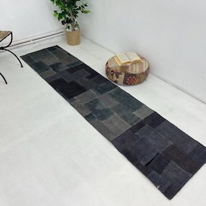 Puede incluir: Una alfombra larga y rectangular de patchwork hecha de tela vaquera en tonos azules y grises. La alfombra está colocada sobre un suelo blanco con un pequeño otomano redondo y estampado y un libro encima.