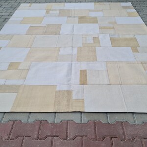 Puede incluir: Una alfombra grande, rectangular y de patchwork con fondo blanco y cuadrados beige. La alfombra está hecha de diferentes materiales y texturas, creando un diseño único e interesante.