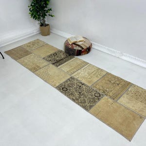 May include: A long, rectangular patchwork rug featuring a mix of beige and brown patterns. The rug is constructed from multiple rectangular sections. It is laid out on a white floor.