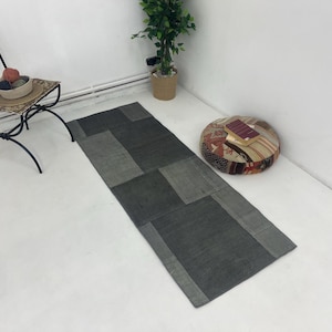 May include: A gray and black patchwork runner rug with a distressed look. The rug is laid out on a white floor with a small potted plant and a colorful patterned floor cushion in the background.