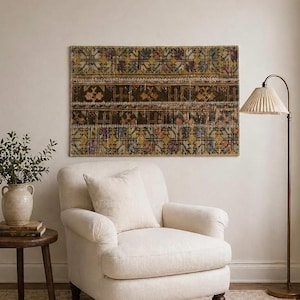 May include: A rectangular, patterned rug with a dark brown central section and colorful geometric designs. The rug is mounted on a neutral wall above a cream-colored armchair. A floor lamp with a pleated shade stands nearby.