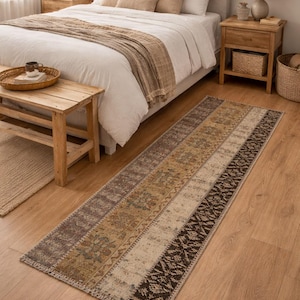 Puede incluir: Una alfombra rectangular larga con un diseño de patchwork en tonos marrones, beige y crema. La alfombra está colocada sobre un suelo de madera en un dormitorio, con una cama y mesitas de noche visibles en el fondo.