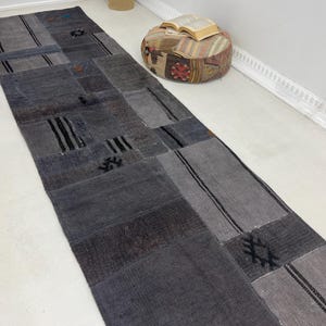 Puede incluir: Una alfombra larga y rectangular de patchwork en tonos grises. La alfombra presenta una variedad de patrones y texturas geométricas, con acentos grises oscuros y negros. La alfombra probablemente está hecha de tela tejida.