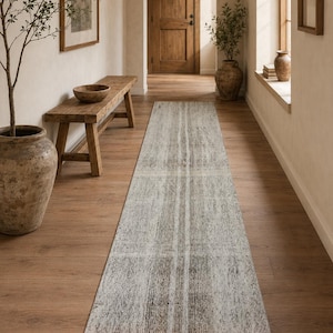 May include: A long, rectangular rug with a distressed, textured appearance. The rug features a neutral color palette with shades of gray, beige, and white, and vertical stripes. The rug is placed on a wooden floor in a hallway setting.