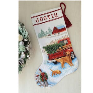 May include: A white Christmas stocking featuring a cross-stitched design of a red pickup truck carrying a pine tree, two golden retriever dogs, a red barn, and the name JUSTIN in red letters. A red tassel hangs from the top corner of the stocking.