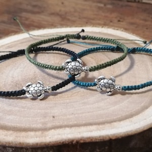 Könnte beinhalten: Drei handgefertigte Armbänder mit Schildkrötenanhängern. Die Armbänder sind schwarz, olivgrün und türkisblau, mit einem geflochtenen Kordeldesign. Jedes Armband hat einen silberfarbenen Schildkrötenanhänger in der Mitte. Auf einer Holzoberfläche präsentiert.