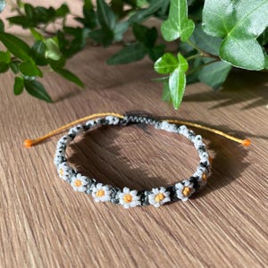 Op de afbeelding: Een zwart-witte vriendschapsband met witte madeliefjes en een gele accent. De armband is gemaakt van een geknoopte touw.