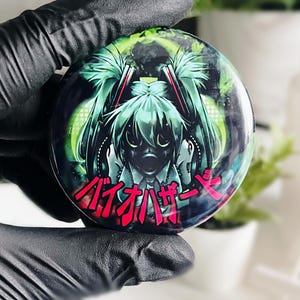 Op de afbeelding: Een ronde knop met een groen, zwart en wit anime-personage met een gasmasker. Het personage heeft lang groen haar en draagt een zwart-witte outfit. De tekst "バ イ オ ハ ザ ー ド" staat in rood op de knop.