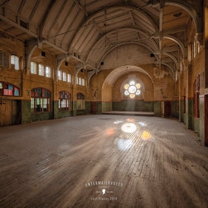 Könnte beinhalten: Ein großer, leerer Raum mit einer hohen gewölbten Decke und Holzböden. Der Raum hat große Fenster mit Buntglas und einer grün gestrichenen Fußleiste. Der Raum wird von Sonnenlicht beleuchtet, das durch die Fenster strömt.