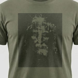 May include: Olive green t-shirt featuring a dark, faded image of a face. The graphic is centered on the chest and has a distressed, vintage appearance. The shirt has short sleeves and a crew neck.