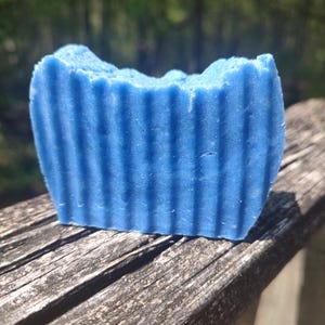 Puede incluir: Una pastilla de jabón azul rectangular con estrías verticales. El jabón está sobre una superficie de madera envejecida, con un fondo verde borroso. El jabón mide unos 8 cm de largo.