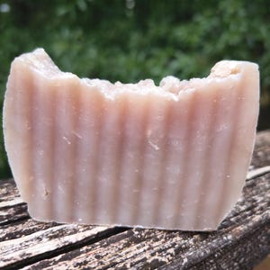 May include: A close-up of a rectangular bar of handmade soap. The soap has a light pink hue with vertical ridges on the sides. The top edge has a rough, uneven texture. The soap is sitting on a weathered wooden surface.