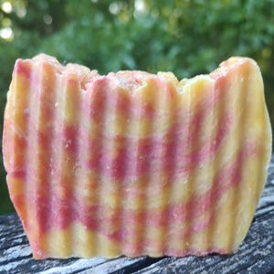 May include: A handmade bar of soap with a textured, ridged surface. The soap features alternating bands of yellow and pink, creating a striped pattern. The soap is rectangular and appears to be freshly cut.