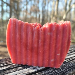 Puede incluir: Una barra rectangular de jabón artesanal de un tono rojo vibrante. El jabón tiene una superficie texturizada y acanalada, y un borde superior ligeramente irregular. La barra descansa sobre una superficie de madera envejecida, con un fondo natural borroso.