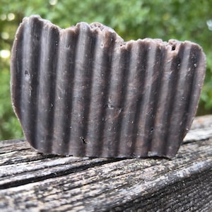 May include: A rectangular bar of dark brown soap with a deeply ribbed, vertical texture on its front surface. The soap has a solid, opaque appearance with subtle variations in the dark brown color throughout its dense, molded structure.