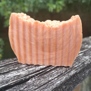Puede incluir: Primer plano de una pastilla de jabón artesanal. El jabón es de color melocotón cálido con estrías verticales en los lados. El borde superior tiene una textura rugosa e irregular. El jabón descansa sobre una superficie de madera envejecida.