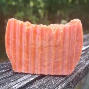 May include: A rectangular bar of orange soap with vertical ridges. The soap is resting on a weathered wooden surface, with a blurred green background of foliage. The soap appears to be handmade.