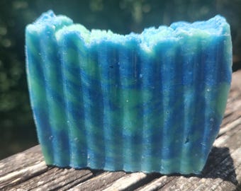 May include: A handmade bar of soap with a blue and green swirled design. The soap has a textured, ridged surface and a slightly uneven top edge. The colors blend together, creating a marbled effect. The soap is sitting on a weathered wooden surface.