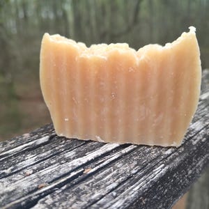 May include: A close-up of a bar of handmade soap with a light brown color and a ridged texture. The soap is sitting on a weathered wooden surface.