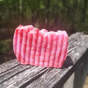 May include: A pink and white striped bar of soap with a slightly uneven edge. The soap is sitting on a weathered wooden surface.