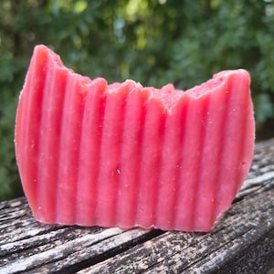 Op de afbeelding: Een rechthoekige roze zeep met verticale ribbels. De zeep, met een onregelmatige bovenrand, ligt op een verweerd houten oppervlak. De achtergrond is een vage weergave van groen.