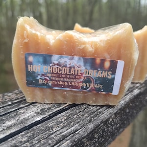 Puede incluir: Una pastilla de jabón marrón claro con una etiqueta que dice "Hot Chocolate Dreams". El jabón está hecho con agua de coco y aceite de oliva y fragancias de primera calidad. El jabón está sobre una superficie de madera desgastada.