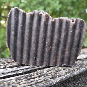 Peut inclure: Un pain de savon brun foncé avec une texture striée. Le savon a un aspect rustique et fait à la main, avec un bord légèrement irrégulier. L'arrière-plan est flou, montrant un feuillage vert et une surface en bois.