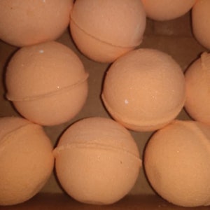 May include: A collection of peach-colored bath bombs, spherical in shape, are arranged in a cardboard container. Each bath bomb is divided into two halves, with a visible seam. The surface has a slightly textured appearance.