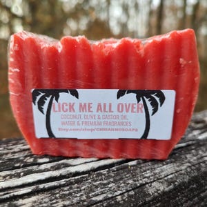 Puede incluir: Una pastilla de jabón roja con una superficie texturizada, envuelta en plástico transparente. El jabón tiene una etiqueta con el texto "LICK ME ALL OVER" y el nombre de la tienda. Ingredientes: coco, oliva y aceite de ricino.
