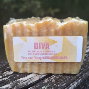 Puede incluir: Una pastilla de jabón rectangular de color amarillo claro. Una etiqueta blanca con la palabra "DIVA" en letras grandes está adherida al frente. La etiqueta también enumera los ingredientes y una dirección web.