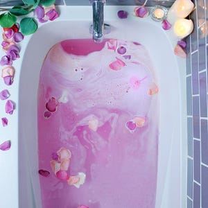 Puede incluir: Una bañera llena de agua rosa y pétalos de rosa. El agua tiene una textura espumosa y está rodeada de azulejos morados. Hojas verdes y más pétalos de rosa se colocan alrededor de la bañera, junto con velas encendidas.