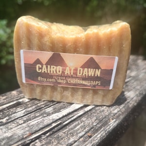 Op de afbeelding: Een stuk zeep met een label waarop "Cairo at Dawn" staat, met daaronder de tekst "Coconut & Olive Oil Base" en "Premium Fragrances". Het label bevat ook de tekst "Etsy.com/shop/CHRISTIANNISOAPS".