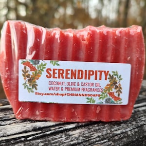 Puede incluir: Una pastilla de jabón roja rectangular envuelta en plástico transparente. El jabón tiene una superficie texturizada y una etiqueta blanca con la palabra "SERENDIPITY" y otro texto. La etiqueta también presenta ilustraciones botánicas.