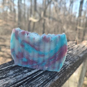 May include: A bar of handmade soap with a swirl pattern in shades of blue, purple, and white. The soap is sitting on a wooden surface.