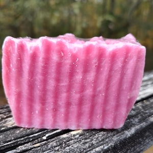 Puede incluir: Una pastilla de jabón rosa rectangular con una textura acanalada. El jabón es de un tono rosa vibrante y parece hecho a mano. El fondo está borroso, lo que sugiere un entorno al aire libre.