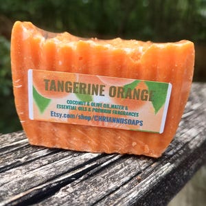 May include: A rectangular bar of orange soap with the words "TANGERINE ORANGE" on a label. The label also includes the words "COCONUT & OLIVE OIL, WATER & ESSENTIAL OILS & PREMIUM FRAGRANCES" and a website address.