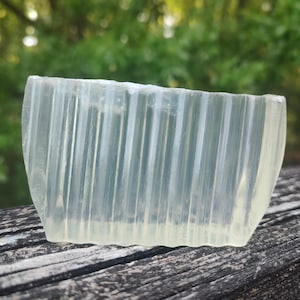 以下が含まれることがあります： 垂直の溝が入った長方形の半透明の石鹸。石鹸は薄い黄色で、風化した木製の表面に置かれています。背景には、ぼやけた緑色の葉があります。